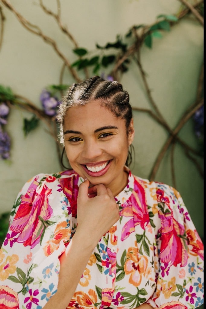 Picture is of a young woman with a big smile with teeth and large Fulani braids wearing a vibrant floral shirt in front of a wall covered in vines