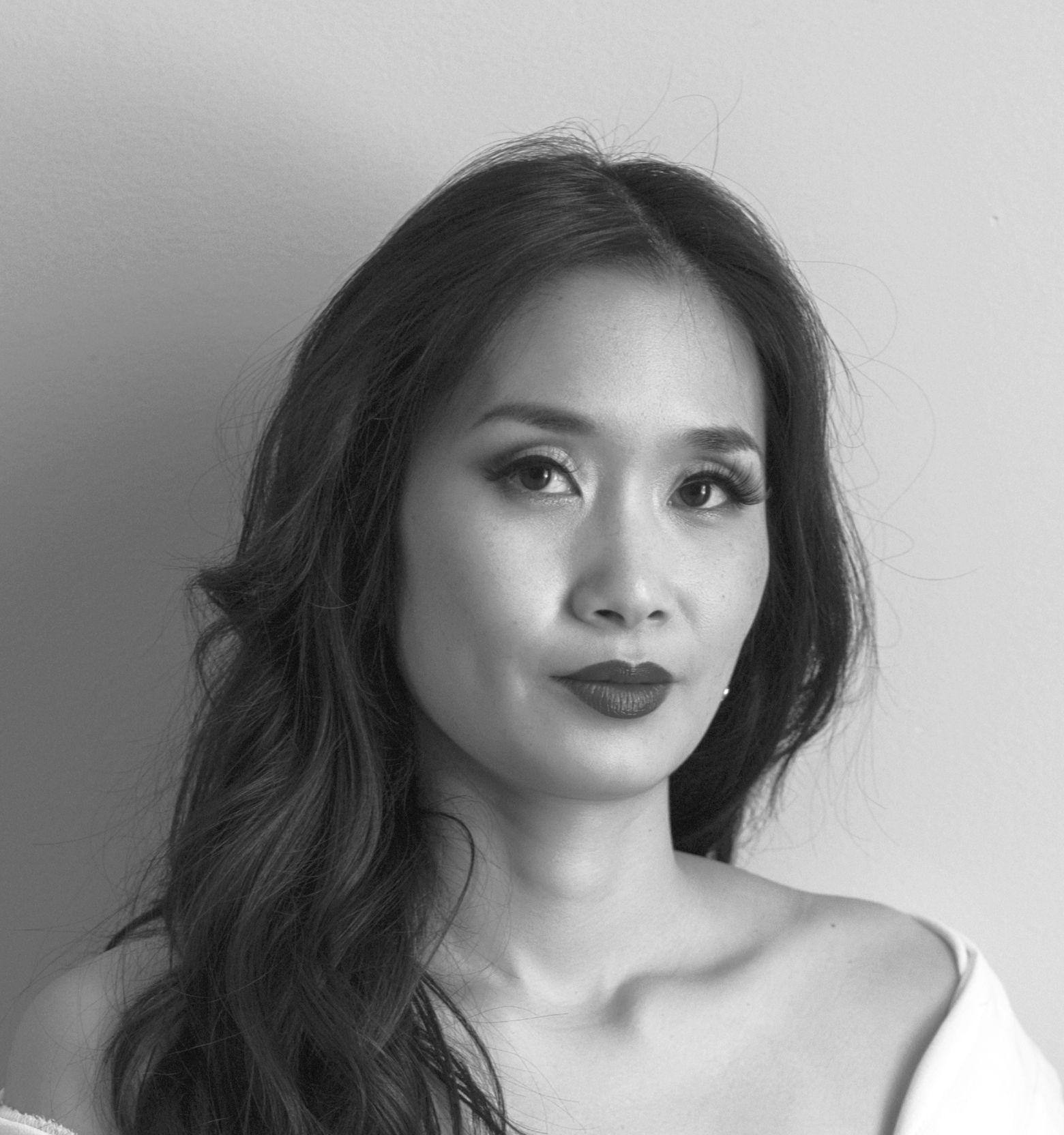 A black and white photographic portrait of a woman with long black hair. She is wearing a white top that covers her shoulders and is looking at the camera with a slight smile.