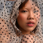 A person with a bleached brow and subtle blush posing with a semi-sheer black and white polka dot fabric framing their face.
